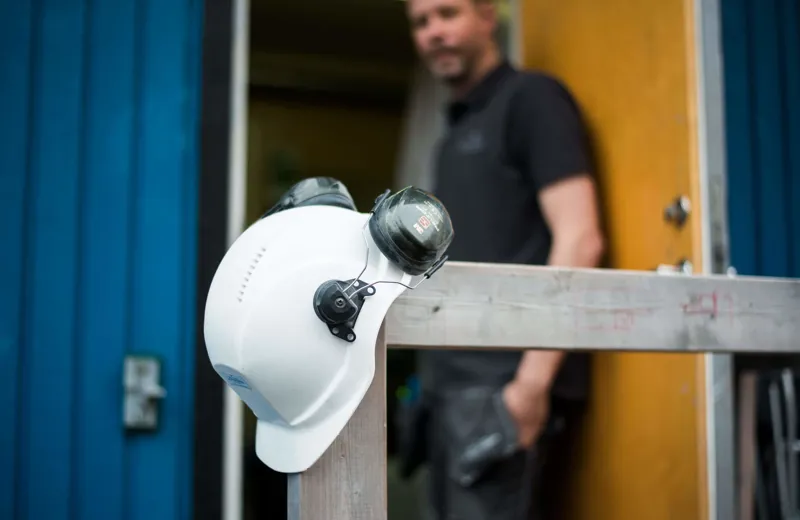 Elektriker i bakgrund, hjälm och hörselkåpor i förgrund.